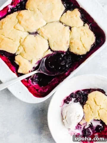 Freshly baked blueberry cobbler in a white baking dish and a scoop of blueberry cobbler in a white bowl with a vanilla scoop of ice cream.