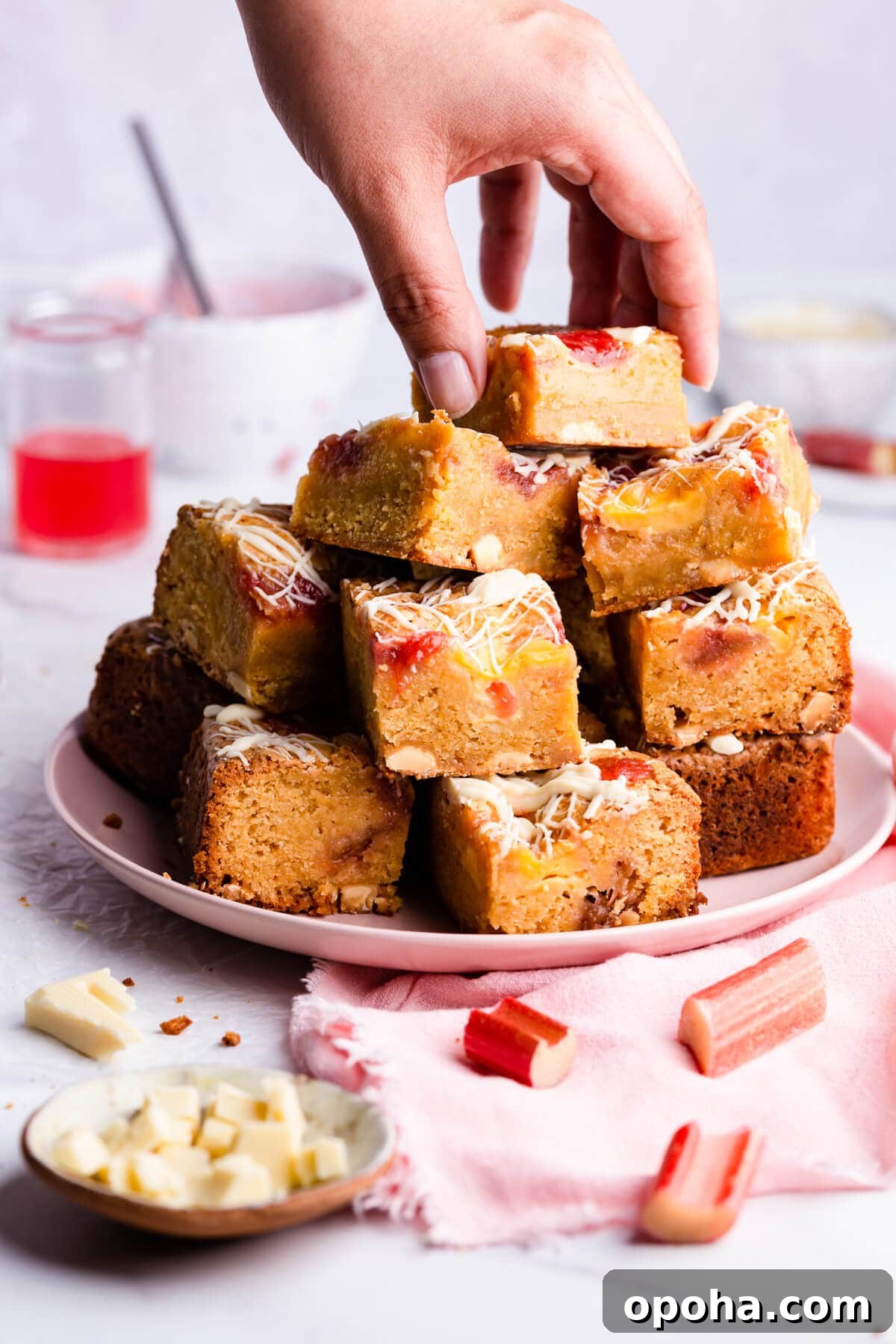 Rhubarb and Custard Blondies Recipe 5 side close up at a plate filled with blondies cake squares and a person taking a piece from the top