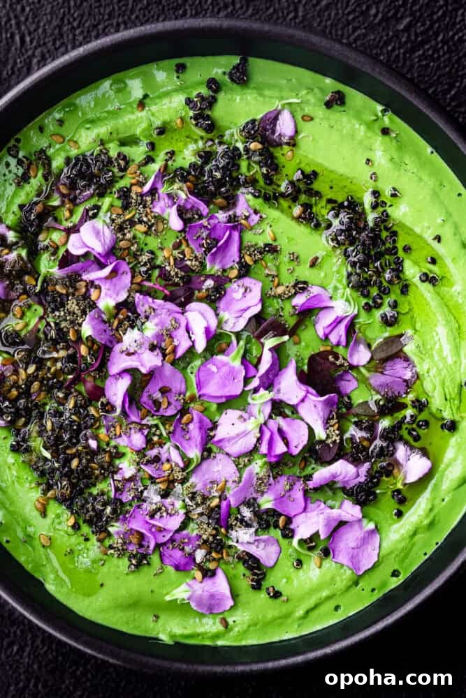 Green tahini topped with nuts, seeds, purple edible flowers, and herbs in a black bowl.