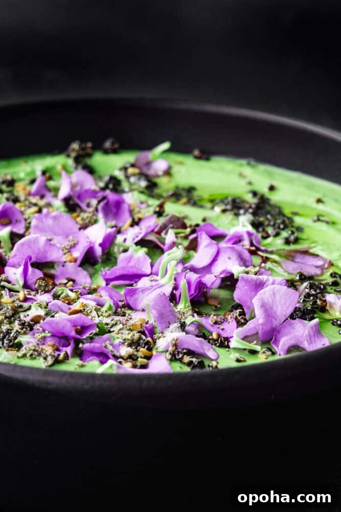 Creamy green tahini sauce topped with purple edible flowers and seeds in a black bowl