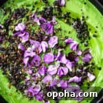 Green tahini topped with nuts, seeds, purple edible flowers, and herbs in a black bowl.