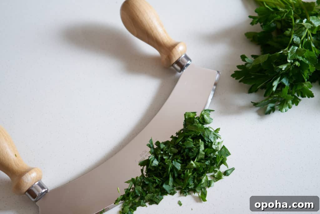 A mezzaluna with wooden handles, chopped parsley, and a bundle of fresh parsley on a white surface.