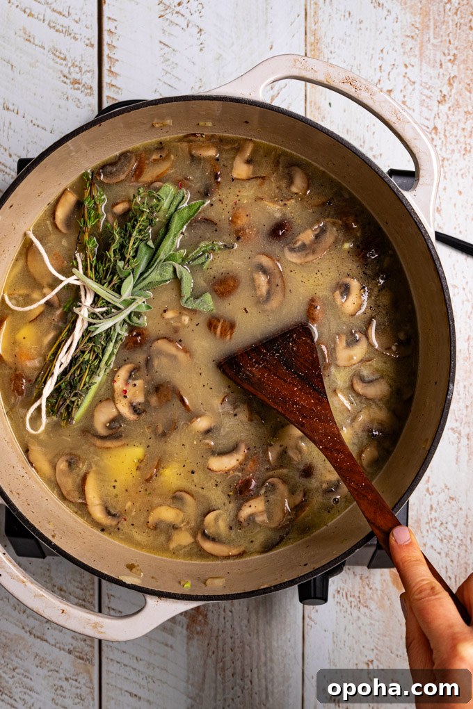 Overhead photo of a white Dutch oven with soup in it. There are lots of mushrooms floating on the surface with a bundle of herbs. A hand is holding a wooden spoon in the soup. The scene has been photographed on a distressed white wood plank surface. #foodphotography