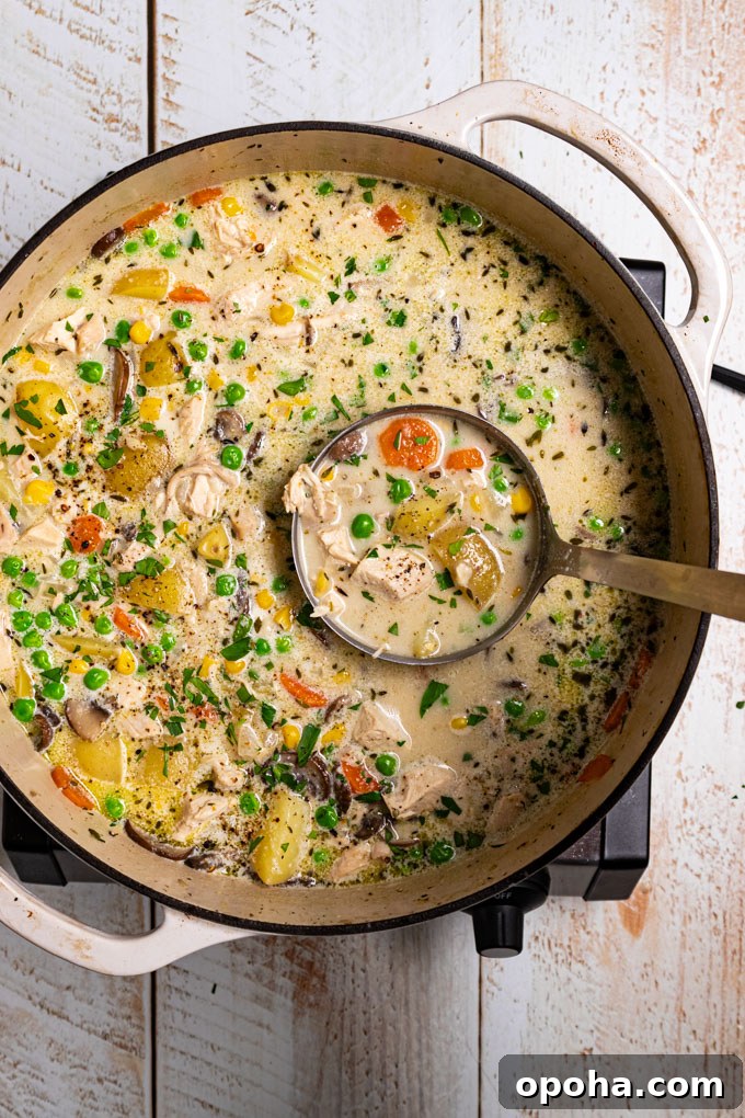 An overhead phot of a white Dutch oven with chicken pot pie soup in it. The soup is creamy with chunks of potatoes, carrots, peas, and mushrooms. It is garnished with fresh parsley and black pepper. A ladle is spooning some of the soup up to the camera. The scene has been photographed on a distressed white wood plank surface. #foodphotography