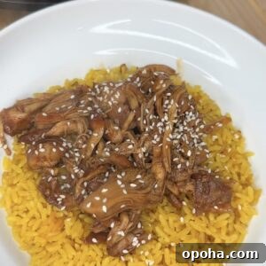 A white bowl filled with yellow rice topped with Crockpot Honey Garlic Chicken in sauce and sprinkled with white sesame seeds.