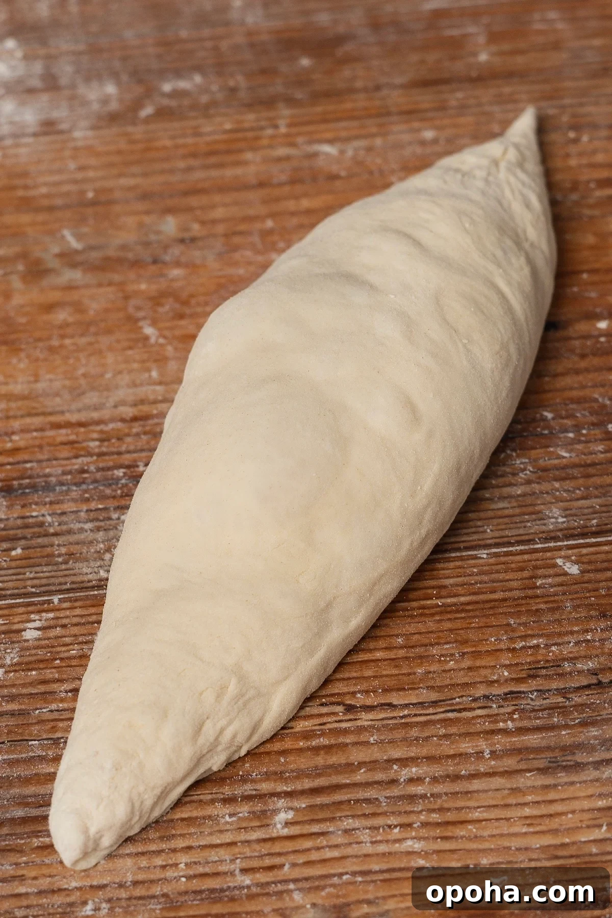 A piece of shaped bread dough rests on a wooden surface, ready for baking. The dough is elongated with tapered ends, resembling a boat shape. Dustings of flour are visible on the surface, enhancing the rustic appearance.