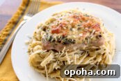 Pesto Parmesan Pork chop served on a bed of linguini pasta on a white plate with a fork on the left sitting on a mustard yellow napkin.
