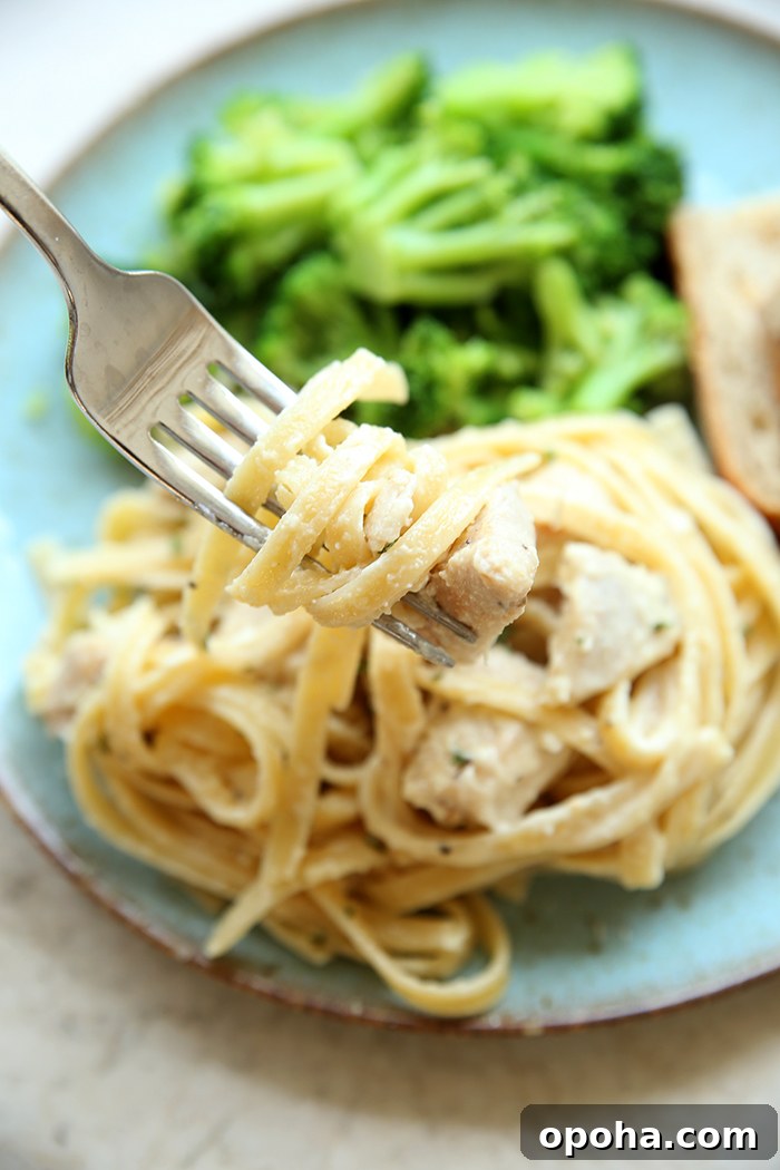 A plate with cooked broccoli, sliced bread, and a scoop of lemon chicken pasta with a fork held above wrapped with noodles and chicken.