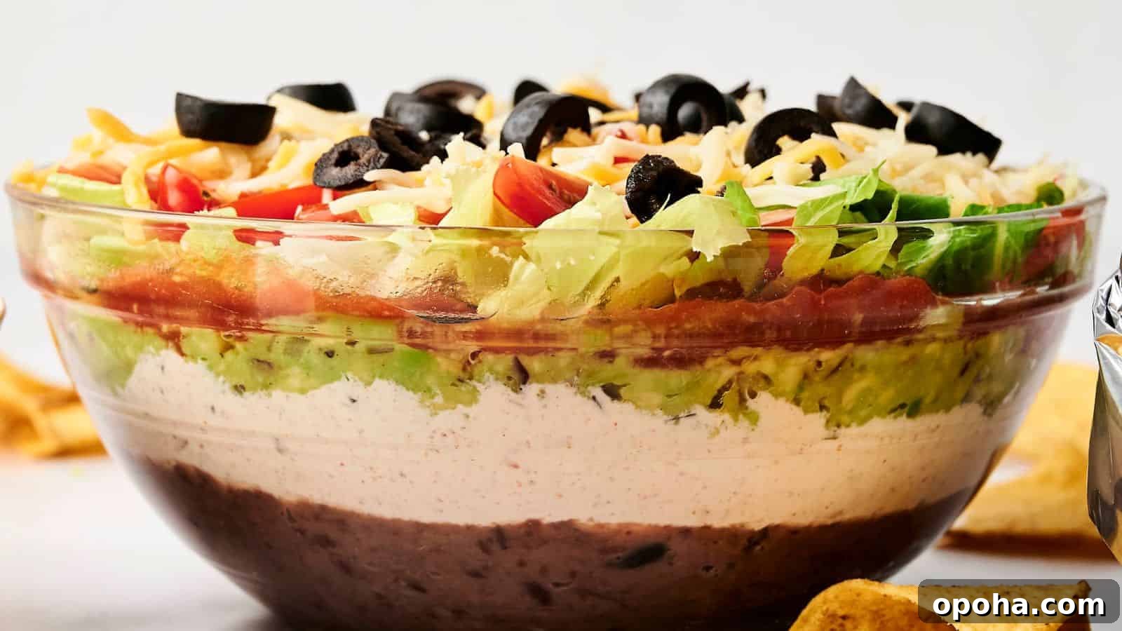 19 Summer Cookout Side Dishes I Make Every Weekend 14 A clear glass bowl with 7 Layer Dip featuring beans, white cream, guacamole, diced tomatoes, shredded cheese, lettuce, and black olives.