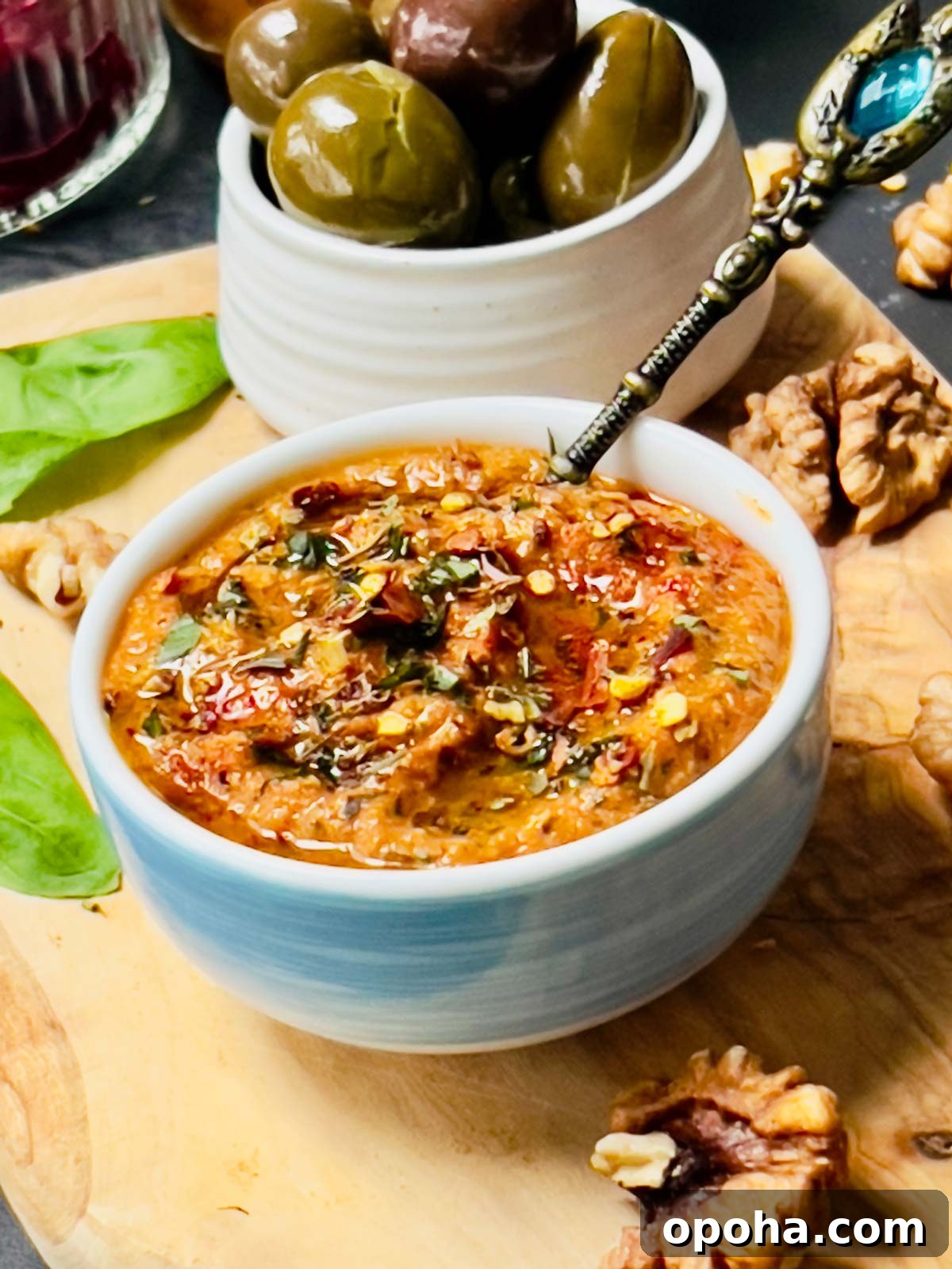 Close up of the pepper sauce with a fancy brass looking teaspoon tucked in and some olives in the background.