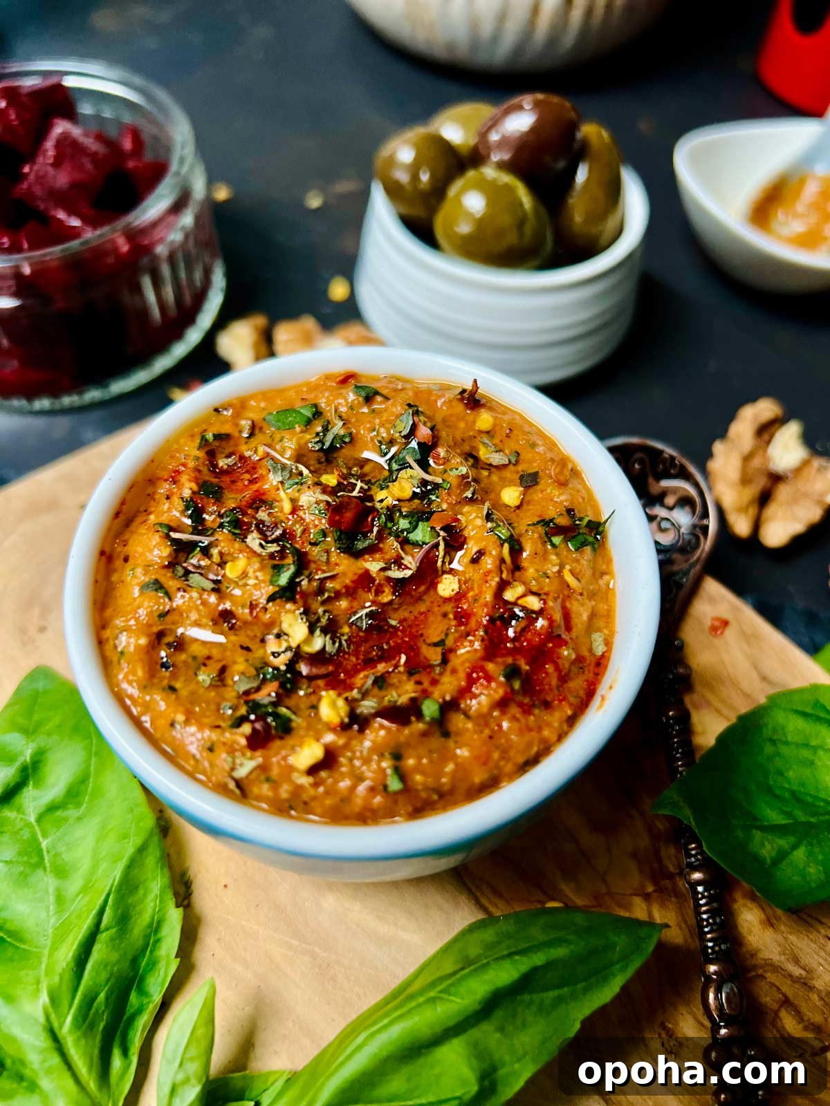 Red bell pepper sauce in a white ceramic dish on a wooden board with basil leaves on and olives and beetroot chunks in the background.
