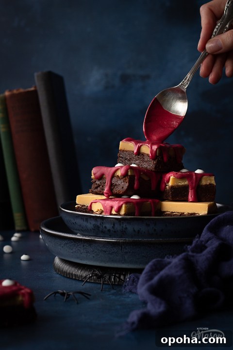 Halloween Millionaire Brownies: Rich Chocolate Caramel Treats 2 Front view of someone pouring melted red chocolate on a pyramid of Halloween Millionaire brownies, set on stacked blue plates against a deep blue hand painted backdrop. There is also a blue linen napkin, fake spiders, edible eyes and some old books in the background.