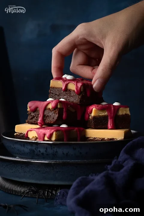 Halloween Millionaire Brownies: Rich Chocolate Caramel Treats 6 Front view close up of someone reaching for a Halloween Millionaire brownie on top of a pyramid of them, set on stacked blue plates against a deep blue hand painted backdrop. There is also a blue linen napkin and some fake spiders in the background.