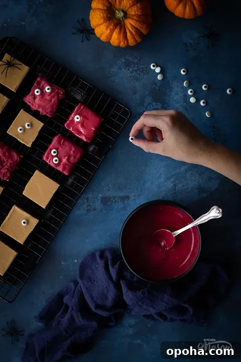 Halloween Millionaire Brownies: Rich Chocolate Caramel Treats 3 Flat lay view of Halloween millionaire brownies on a cooling rack being decorated with edible eyes. There is also a bowl of red white chocolate in a black bowl, a blue linen napkin, 2 small pumpkins and some edible eyes in the background. Set on a deep blue hand painted backdrop.