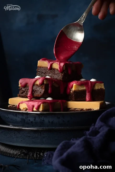 Halloween Millionaire Brownies: Rich Chocolate Caramel Treats 11 Front view close up of someone pouring melted red chocolate on a pyramid of Halloween Millionaire brownies, set on stacked blue plates against a deep blue hand painted backdrop. There is also a blue linen napkin and some fake spiders in the background.