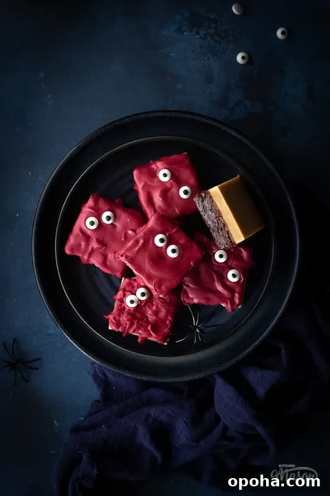 Halloween Millionaire Brownies: Rich Chocolate Caramel Treats 12 Flat lay close up of a dark blue plate filled with Halloween millionaire brownies set on a deep blue hand painted backdrop. There are also edible eyes, fake spiders and a blue linen napkin the background.