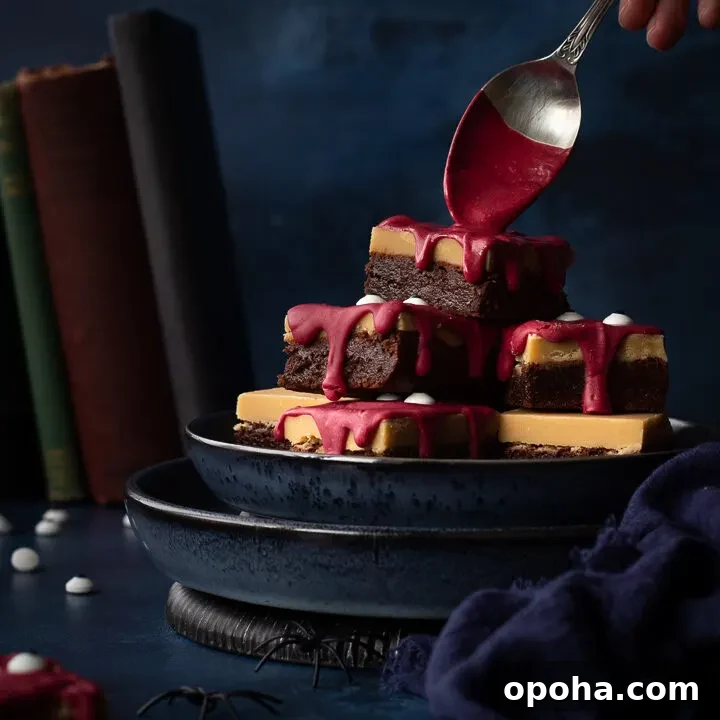 Front view of someone pouring melted red chocolate on a pyramid of Halloween Millionaire brownies, set on stacked blue plates against a deep blue hand painted backdrop. There is also a blue linen napkin, fake spiders, edible eyes and some old books in the background.