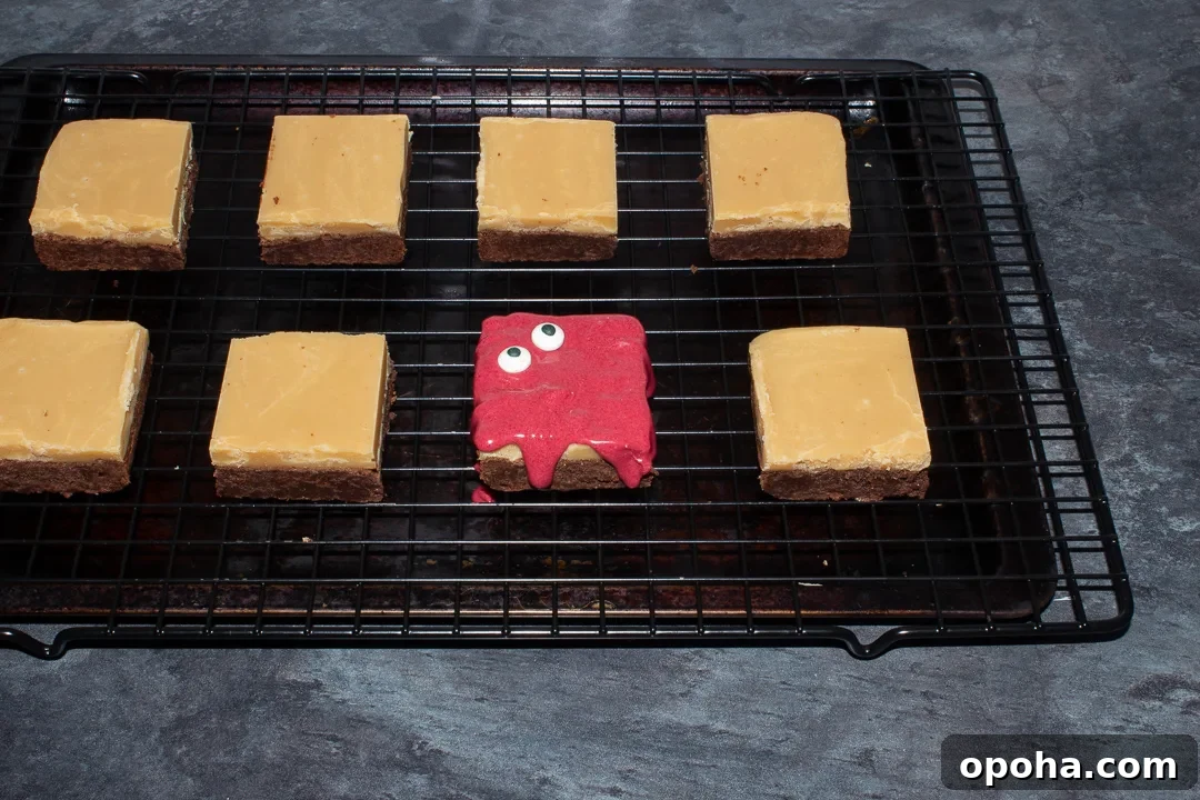 Halloween Millionaire Brownies: Rich Chocolate Caramel Treats 21 Halloween millionaire brownies on a cooling rack set over a baking tray on a kitchen worktop. One has been covered in melted red white chocolate and topped wth 2 edible eyes.