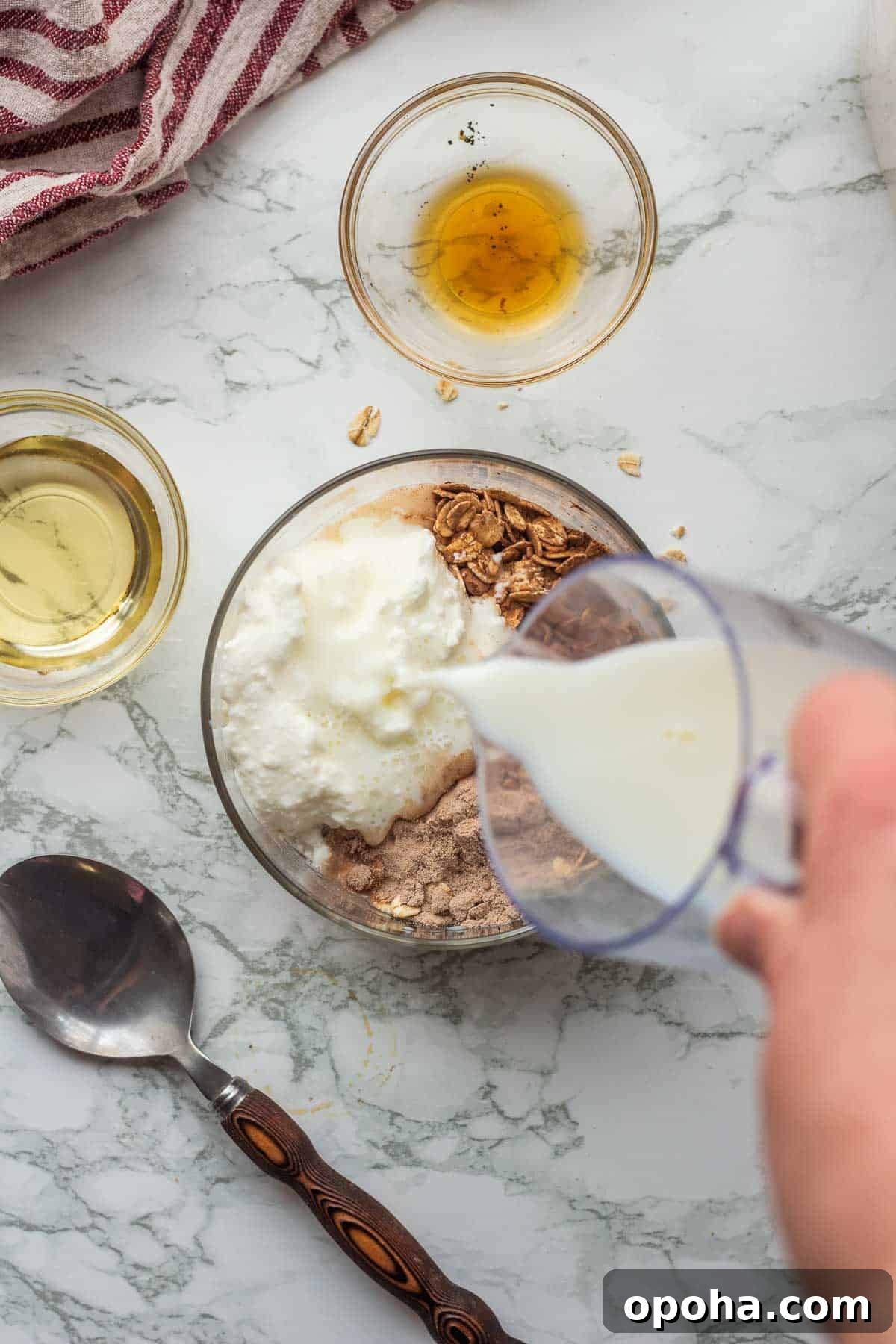 Pouring milk into glass bowl with oats, cocoa powder, protein powder, and greek yogurt.