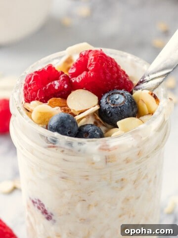 Jar of vanilla overnight oats topped almonds, rapsberries and blueberries with metal spoon.