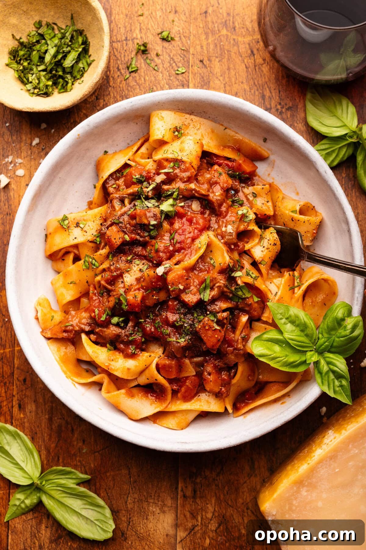 Braised Short Rib Ragu with Pappardelle Pasta 7 A white bowl with short rib beef ragu and pappardelle pasta in it. Around the bowl is fresh basil leaves, a glass of red wine, and a small dish with chopped fresh basil in it all decorated on a wooden background.
