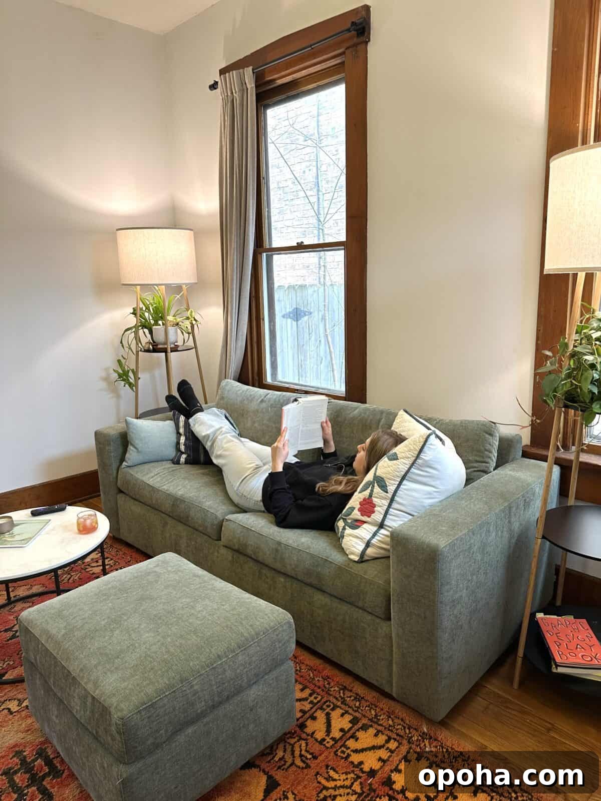 A woman lounges on a green sofa reading a book in a cozy living room with warm lighting, houseplants, and a patterned rug.