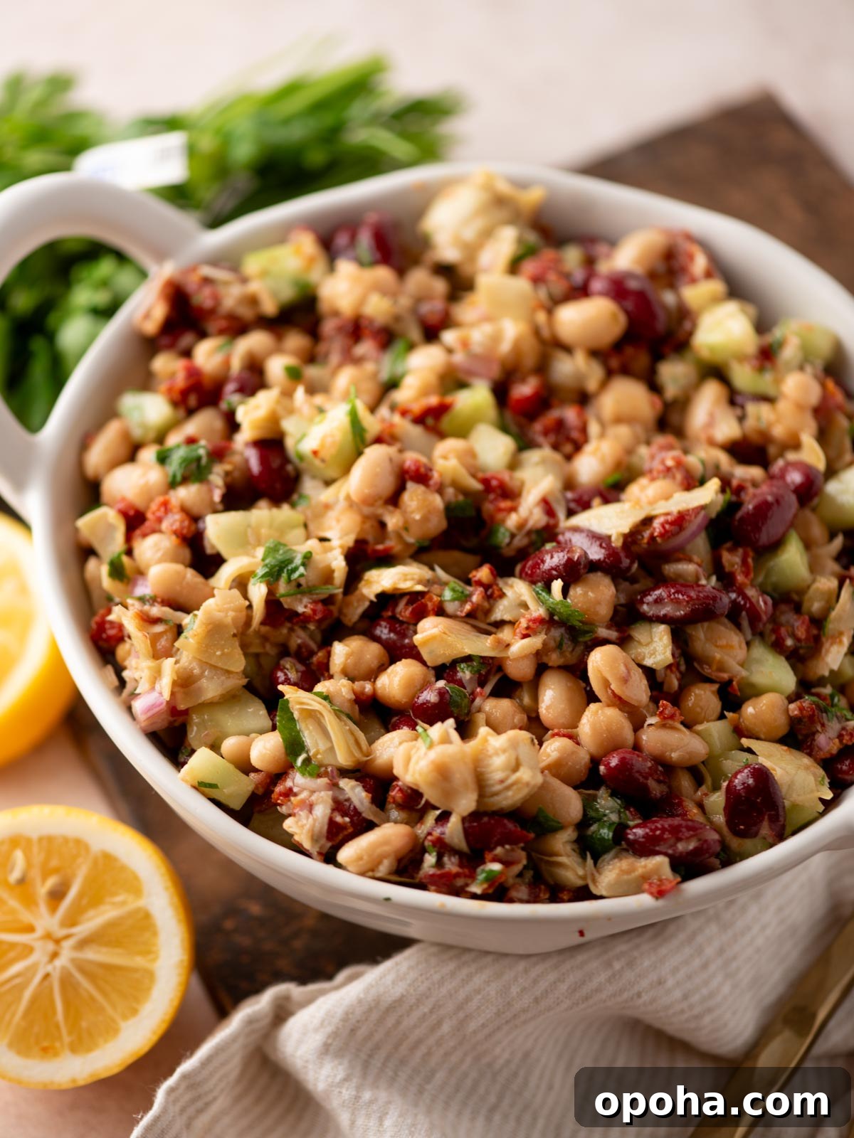 Zesty Three-Bean Salad Recipe for Picnic & Meal Prep 2 A white bowl filled with a colorful bean salad containing kidney beans, chickpeas, artichoke hearts, chopped cucumbers, herbs, and sun-dried tomatoes, with lemon halves and fresh parsley nearby on a wooden surface.