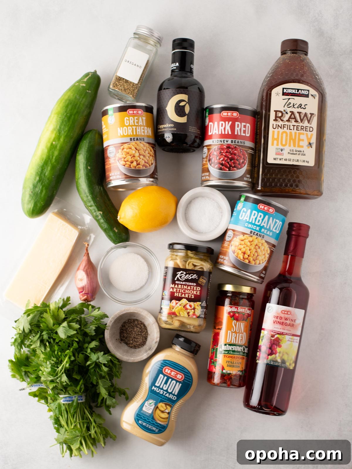 Zesty Three-Bean Salad Recipe for Picnic & Meal Prep 3 A flat lay of various ingredients, including canned beans, cucumber, lemon, honey, red wine vinegar, Dijon mustard, herbs, sun-dried tomatoes, cheese, black pepper, salt, olives, and a small bottle of balsamic vinegar.