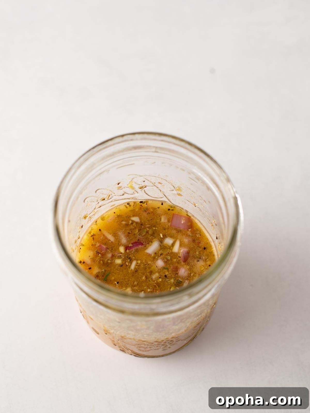 Zesty Three-Bean Salad Recipe for Picnic & Meal Prep 4 A glass jar filled with a chunky vinaigrette-style dressing containing visible bits of red onion and herbs, set against a plain white background.
