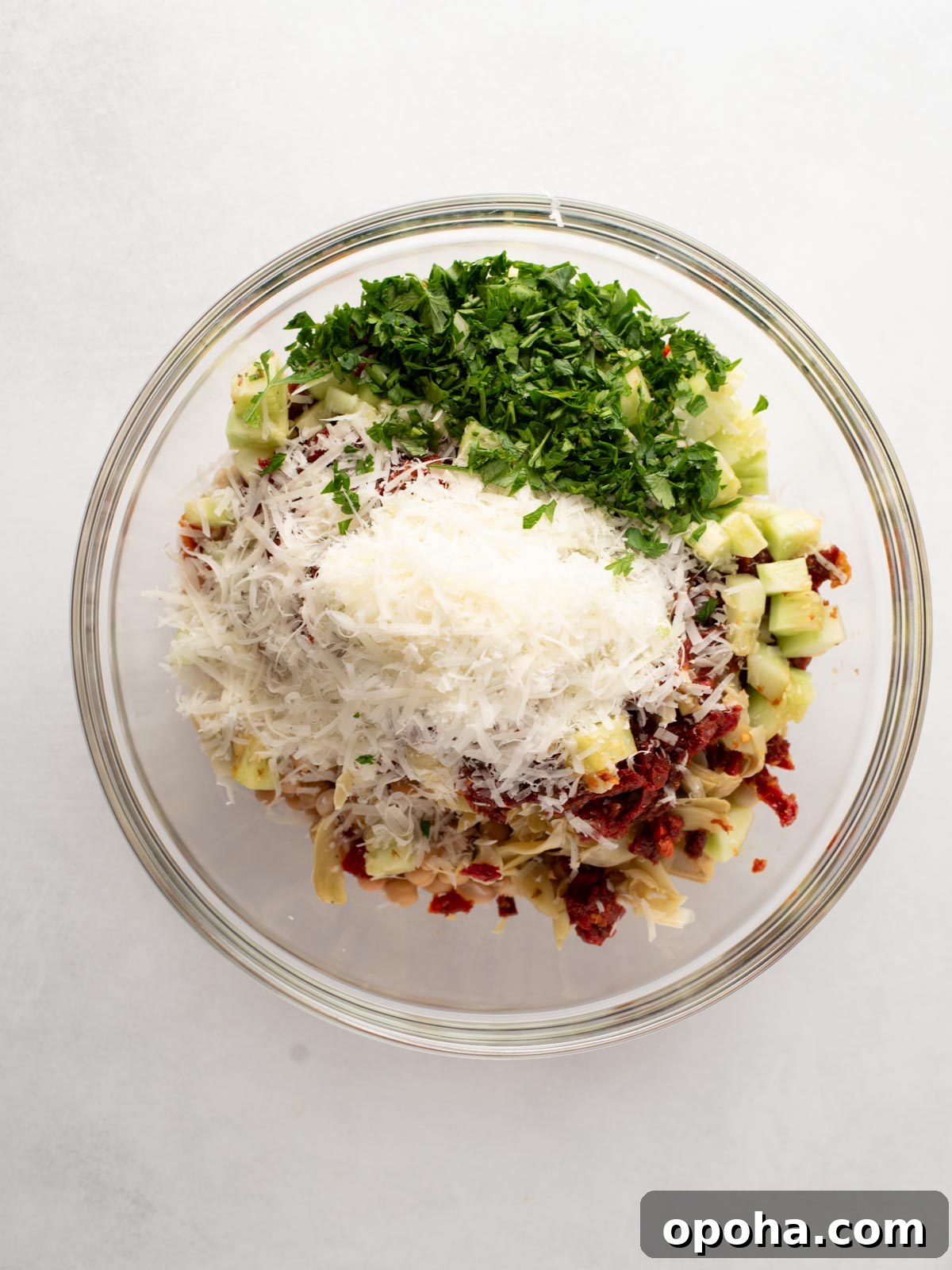 Zesty Three-Bean Salad Recipe for Picnic & Meal Prep 5 A glass bowl filled with chopped parsley, grated cheese, diced celery, sun-dried tomatoes, and artichoke hearts on a white background.