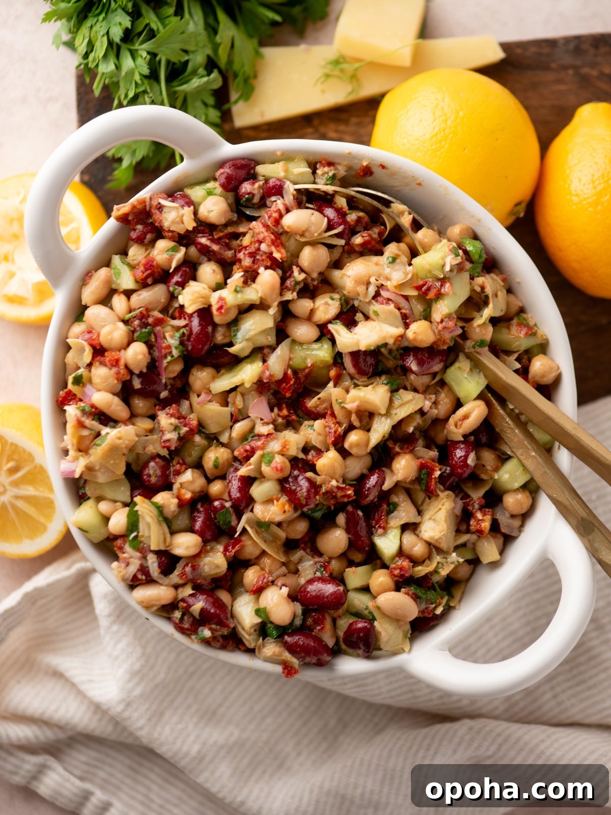 Zesty Three-Bean Salad Recipe for Picnic & Meal Prep 7 A white bowl filled with a colorful bean salad featuring kidney beans, chickpeas, white beans, chopped vegetables, and herbs. Lemons, cheese slices, and fresh parsley are on a wooden board in the background.
