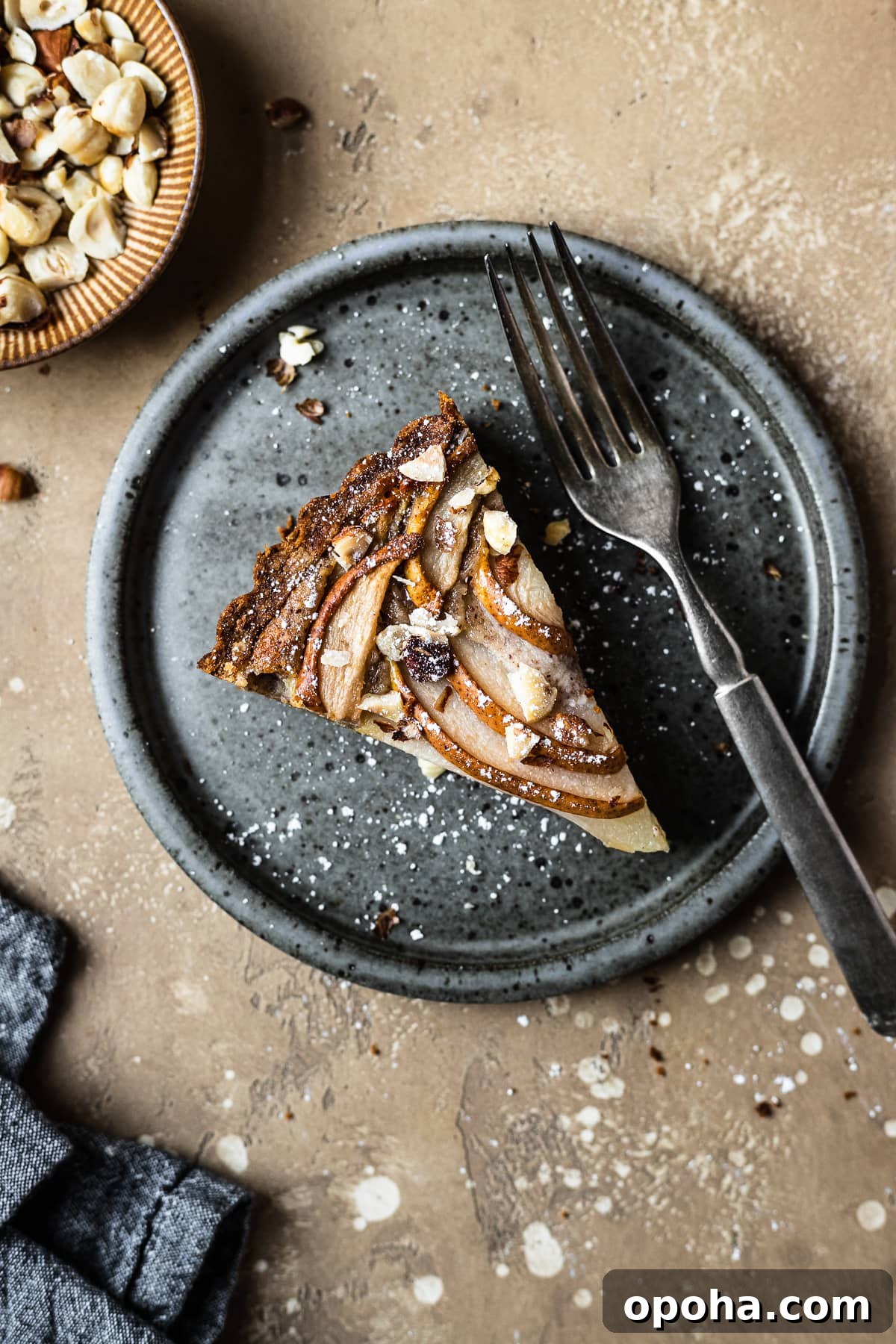 Pear Tart with Hazelnut Frangipane Recipe 2 Pear hazelnut tart slice on a speckled grey blue ceramic plate. A vintage fork rests on the right side of the plate. The plate is on a warm tan textured stone background. A small bit of a textured grey napkin peeks out from the bottom left corner, and a small view of a bowl of chopped hazelnuts is visible at top left.