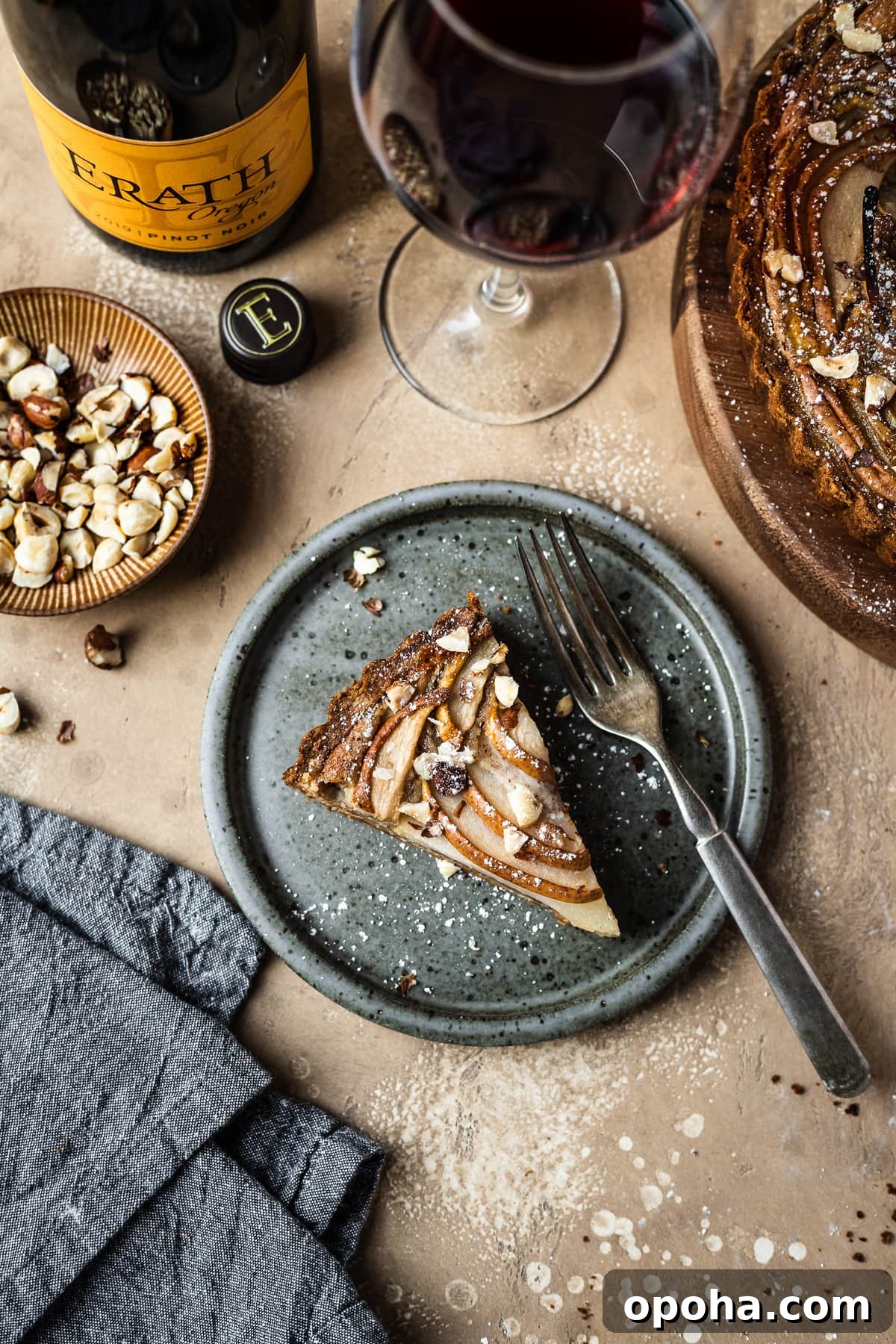 Pear Tart with Hazelnut Frangipane Recipe 12 Pear hazelnut tart slice on a speckled grey blue ceramic plate. A vintage fork rests on the right side of the plate. The plate is on a warm tan textured stone background. A textured grey napkin is at the bottom left corner, and a small bowl of chopped hazelnuts is visible at top left along with a wine bottle and a glass of red wine at top. The remainder of the tart peeks out at top right.
