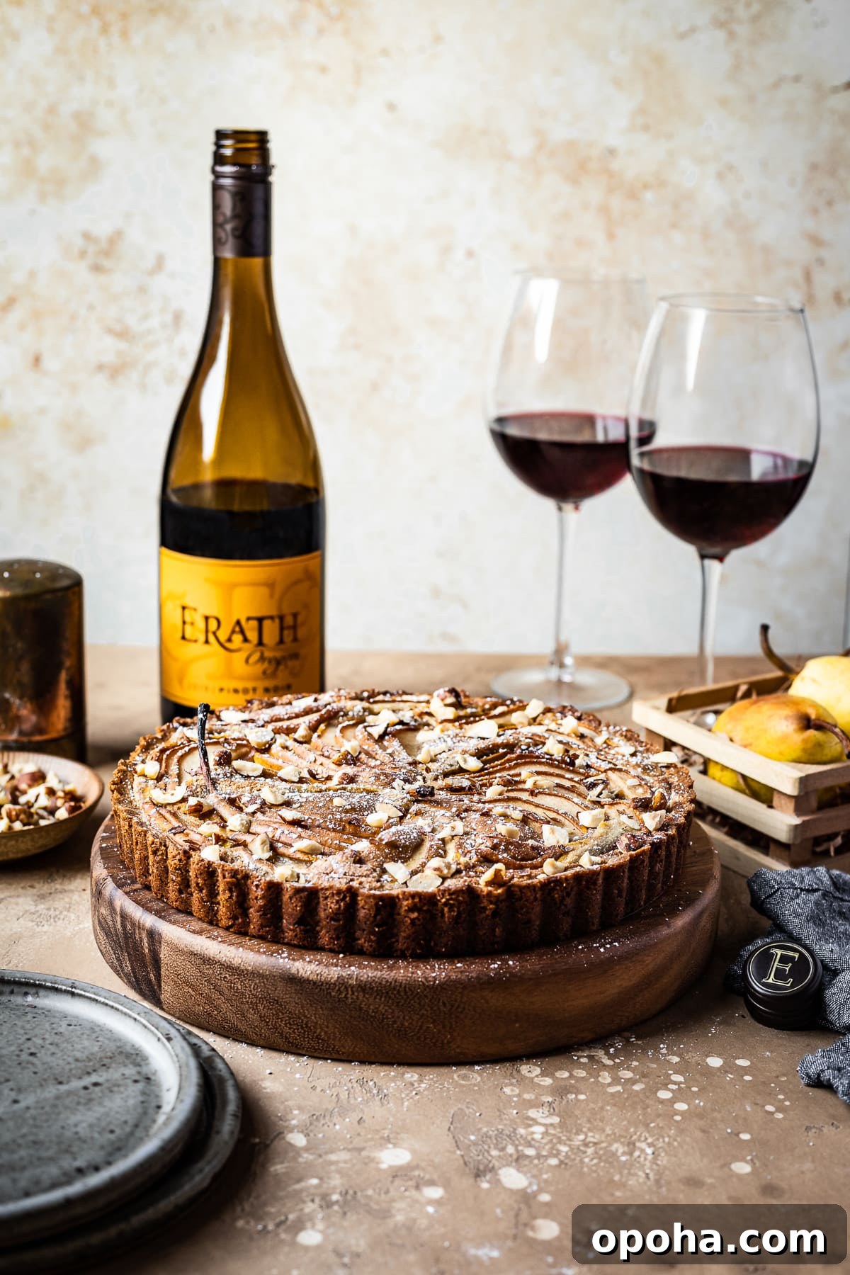 Pear Tart with Hazelnut Frangipane Recipe 3 A front view of a pear tart on a round wooden platter on a textured warm tan background. The tart is surrounded by blue grey ceramic plates, an opened bottle of red wine, two half full glasses of red wine, a small wooden crate of pears, and a textured grey napkin.