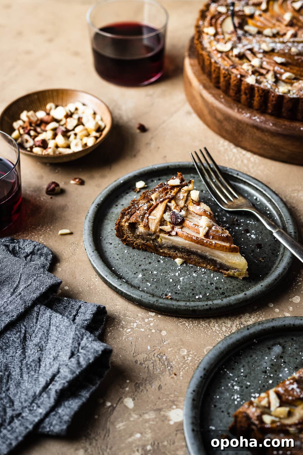 Pear Tart with Hazelnut Frangipane Recipe 4 An angled view of two slices of tart on speckled grey blue ceramic plates. Nearby is the remainder of the tart, a small bowl of hazelnuts, and two stemless wine glasses filled with red wine. A grey textured linen napkin is at bottom left. The background of the image is a warm tan speckled stone.