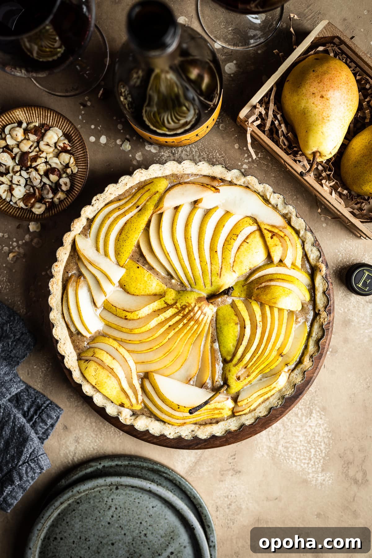 Pear Tart with Hazelnut Frangipane Recipe 5 An unbaked pear hazelnut tart in a round pan is at the center of the image. Surrounding the tart are speckled blue ceramic plates, a bowl of chopped hazelnuts, pears in a wooden crate, a linen napkin, and a bottle of red wine with two filled wine glasses. Everything rests on a textured tan background.