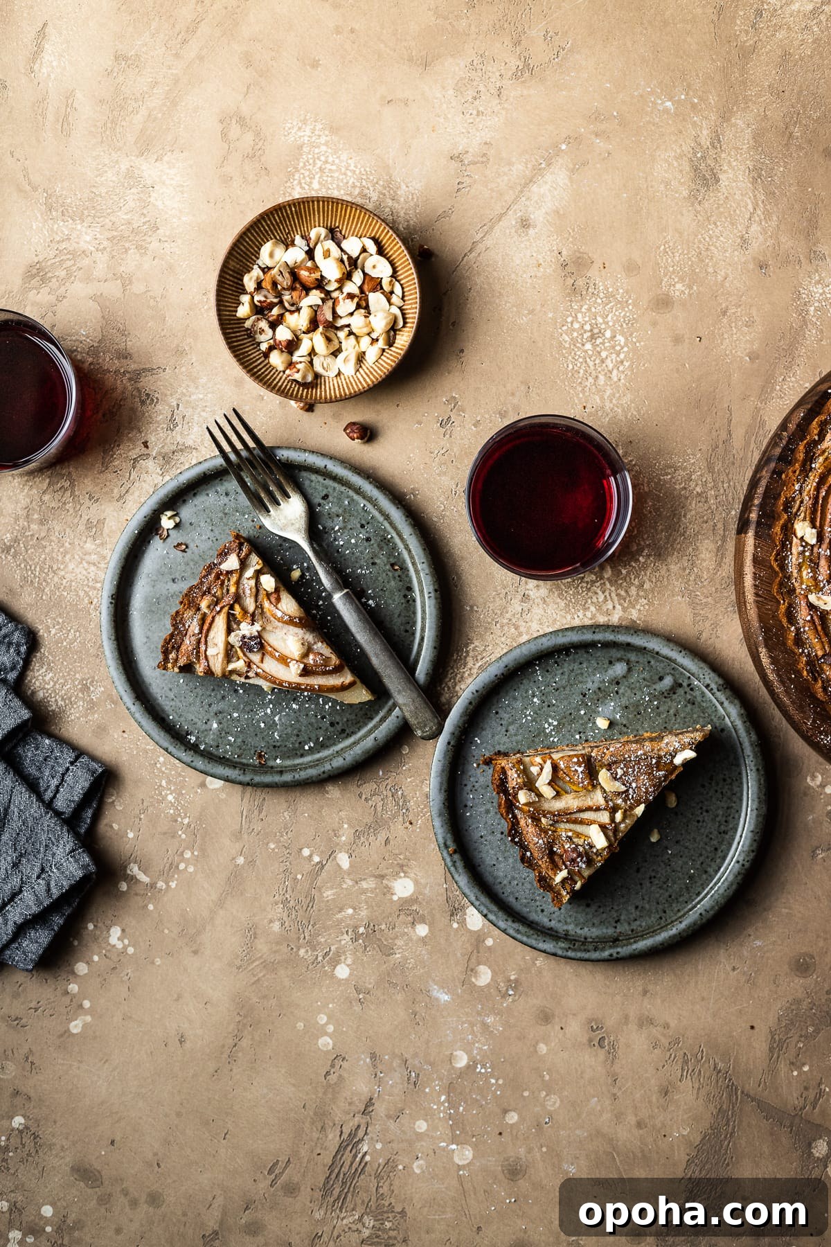 Pear Tart with Hazelnut Frangipane Recipe 7 Two slices of tart on speckled grey blue ceramic plates. To the right is the remainder of the tart. A small bowl of hazelnuts, and two stemless wine glasses filled with red wine surround the plates. A grey textured linen napkin is at lower left. The background of the image is a warm tan speckled stone.