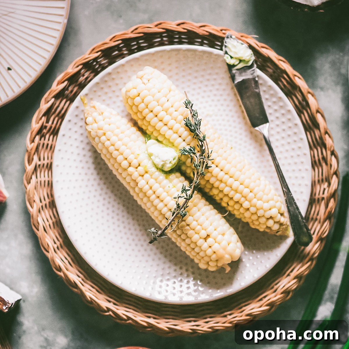 two cobs of plump white corn boiled in coconut milk on a plate with a dollop of spicy chive butter garnished with thyme