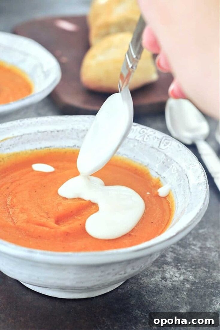 A hand is holding a spoon with bright white cashew cream, adding it to the top of a bowl of bright orange sweet potato soup.