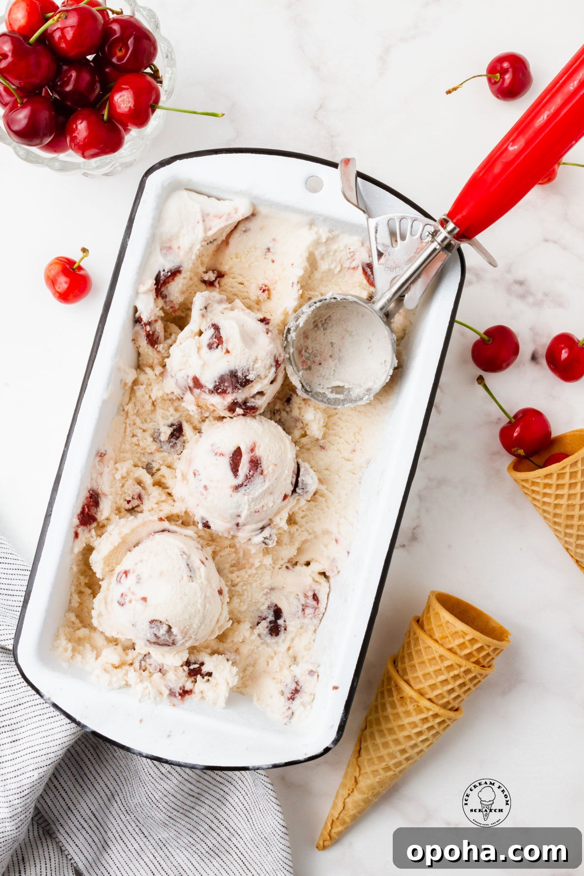 Cherry Gelato Recipe: Creamy Homemade Gelato with Fresh Cherries 2 A white loaf pan filled with cherry gelato. A red and metal ice cream scoop is in the pan, and the ice cream has been scooped. Around the pan are fresh cherries and sugar cones.
