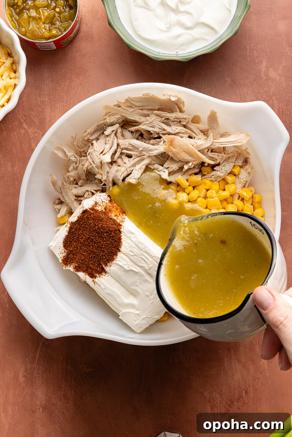 A person's hand pouring green enchilada sauce into a large bowl filled with ingredients to make a green chile chicken enchilada dip