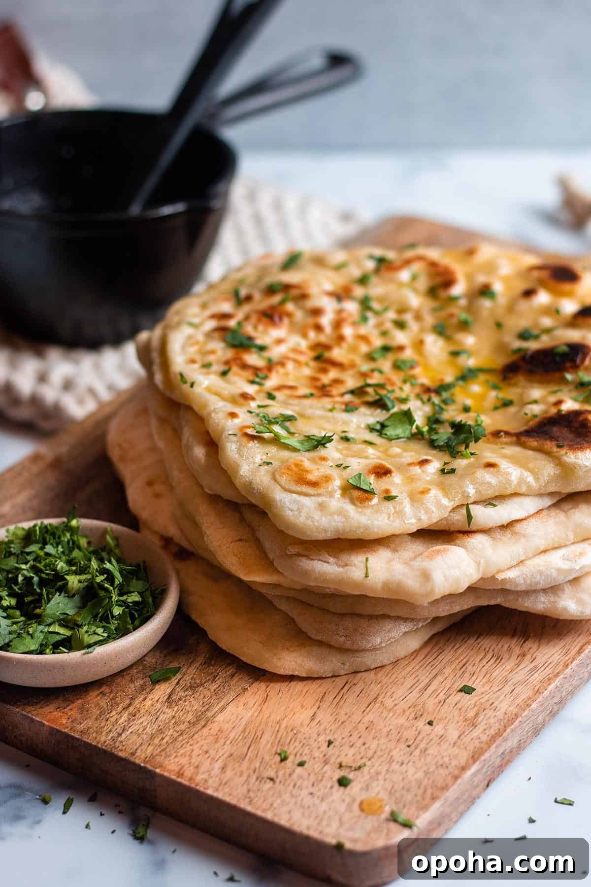 Sourdough Discard Naan Recipe: Crispy, Chewy Flatbread 2 A stack of sourdough naan on a wooden board with cilantro and melted butter. Bowl with fresh cilantro and cast iron butter warmer.