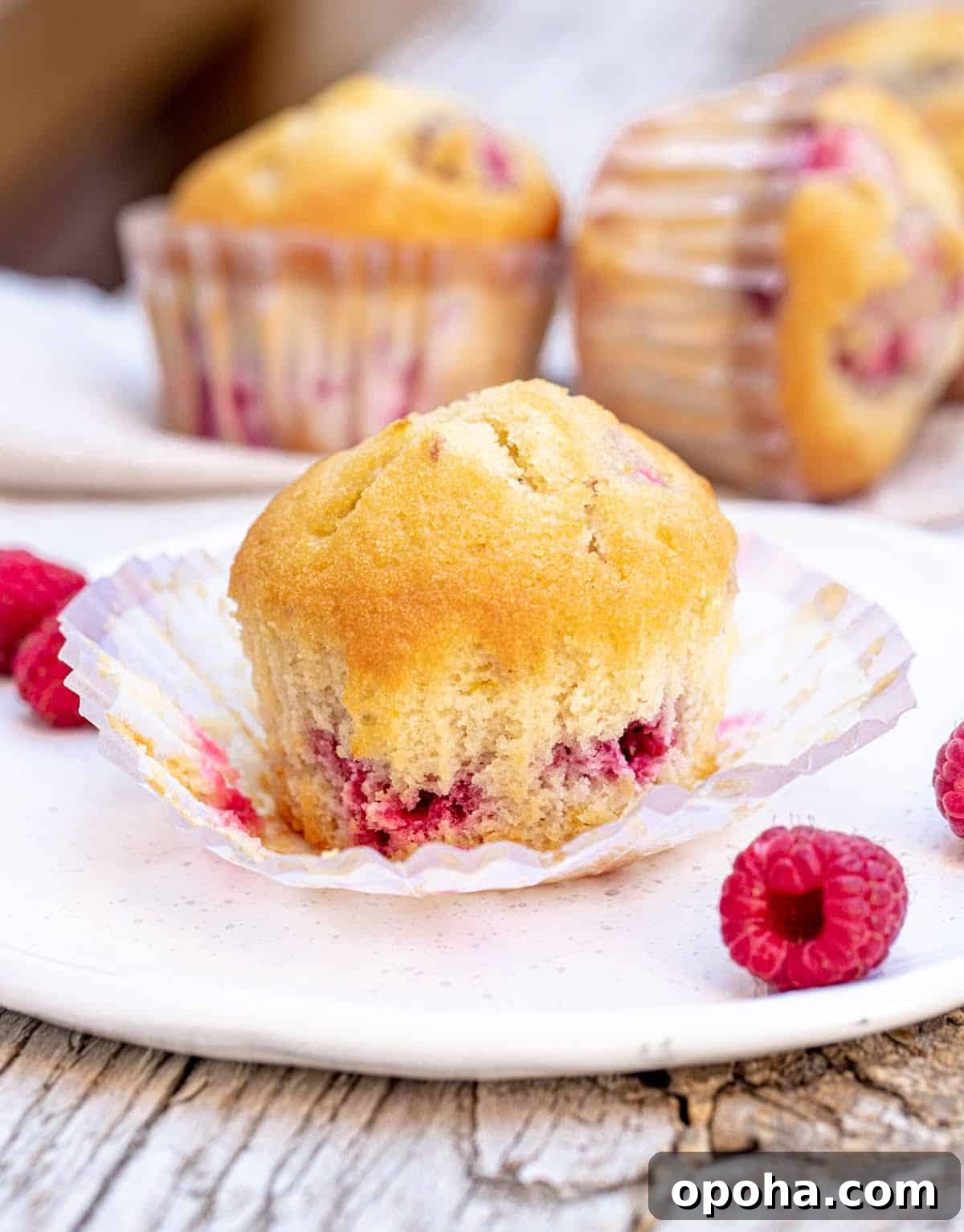Quick Raspberry Muffins Recipe — Light, Tangy, Perfect for Brunch 11 Whole raspberry muffin on opened paper liner on white plate, more muffins in background.