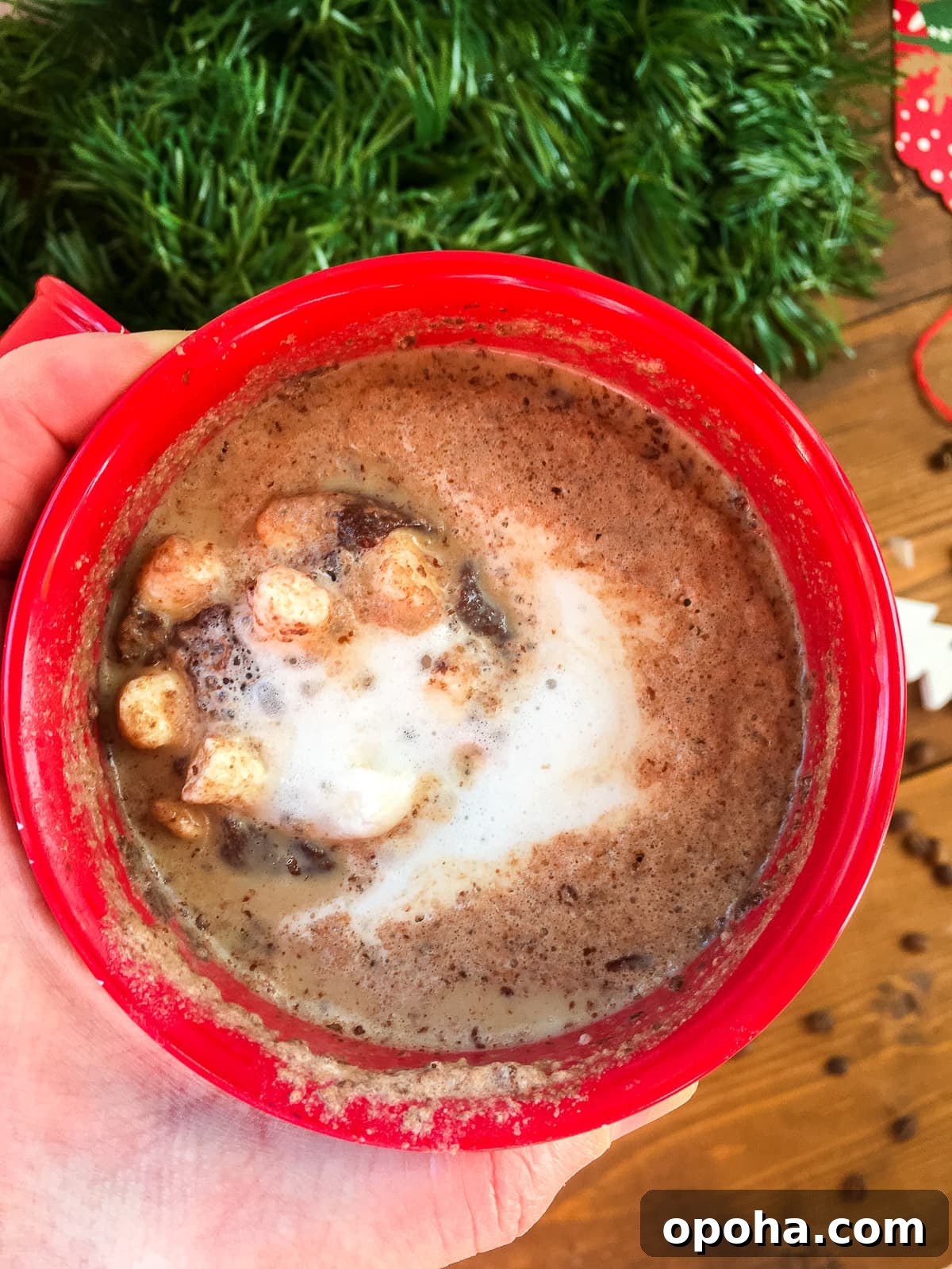 Homemade Mocha Coffee Bombs: Decadent Hot Chocolate Surprise 7 A zoomed out image of a hand holding a red mug of hot cocoa with green garland in the background.