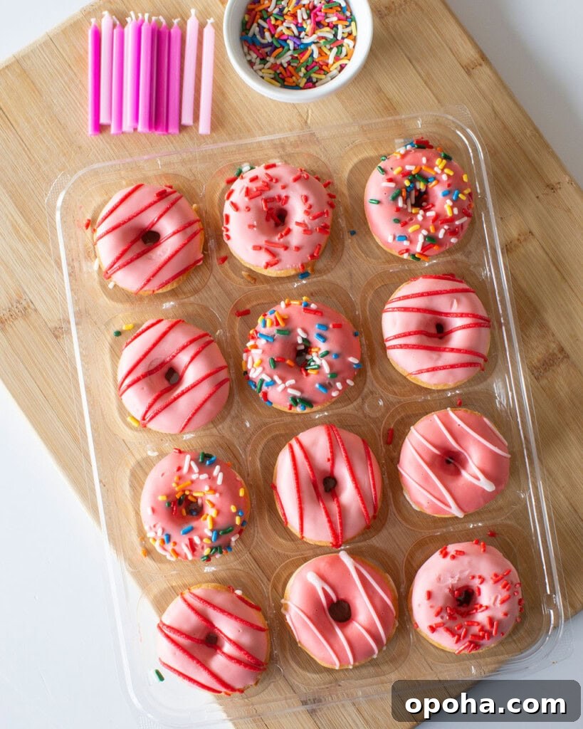Donut Cake Tower Ideas: Show-Stopping Dessert Centerpieces 3 A dozen pink frosted donuts with various sprinkle decorations are arranged in a clear plastic tray. They are placed on a wooden surface next to a small bowl of colorful sprinkles and a group of pink birthday candles.