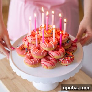 Donut Cake Tower Ideas: Show-Stopping Dessert Centerpieces 5 A person holds a cake stand with pink-frosted donuts topped with colorful sprinkles and lit pink candles. A small bowl of extra sprinkles is on the table. The person wears a pink apron.