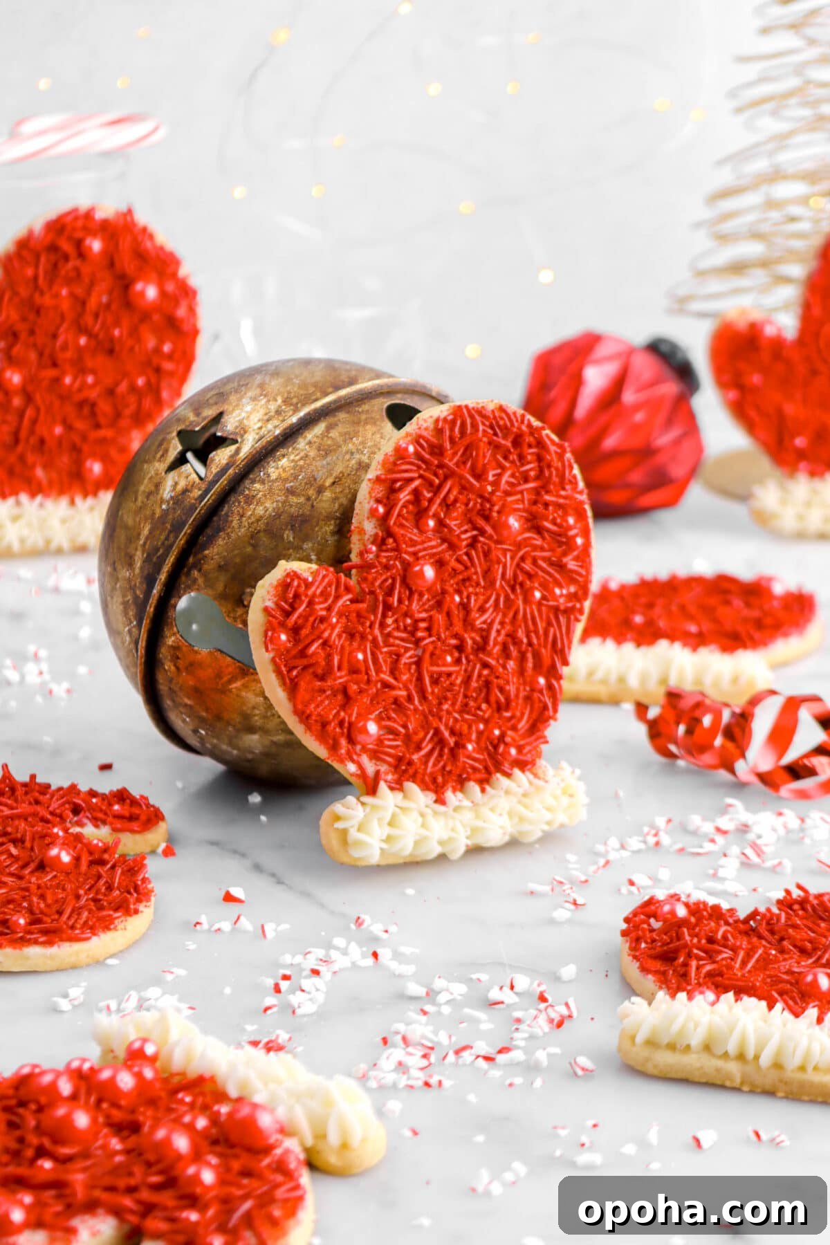 Santa’s Glove Peppermint Sugar Cookies: Festive Holiday Recipe 2 glove shaped sugar cookie propped against large bell, with six more cookies around, red ribbon beside, a red ornament, and fairy lights behind.