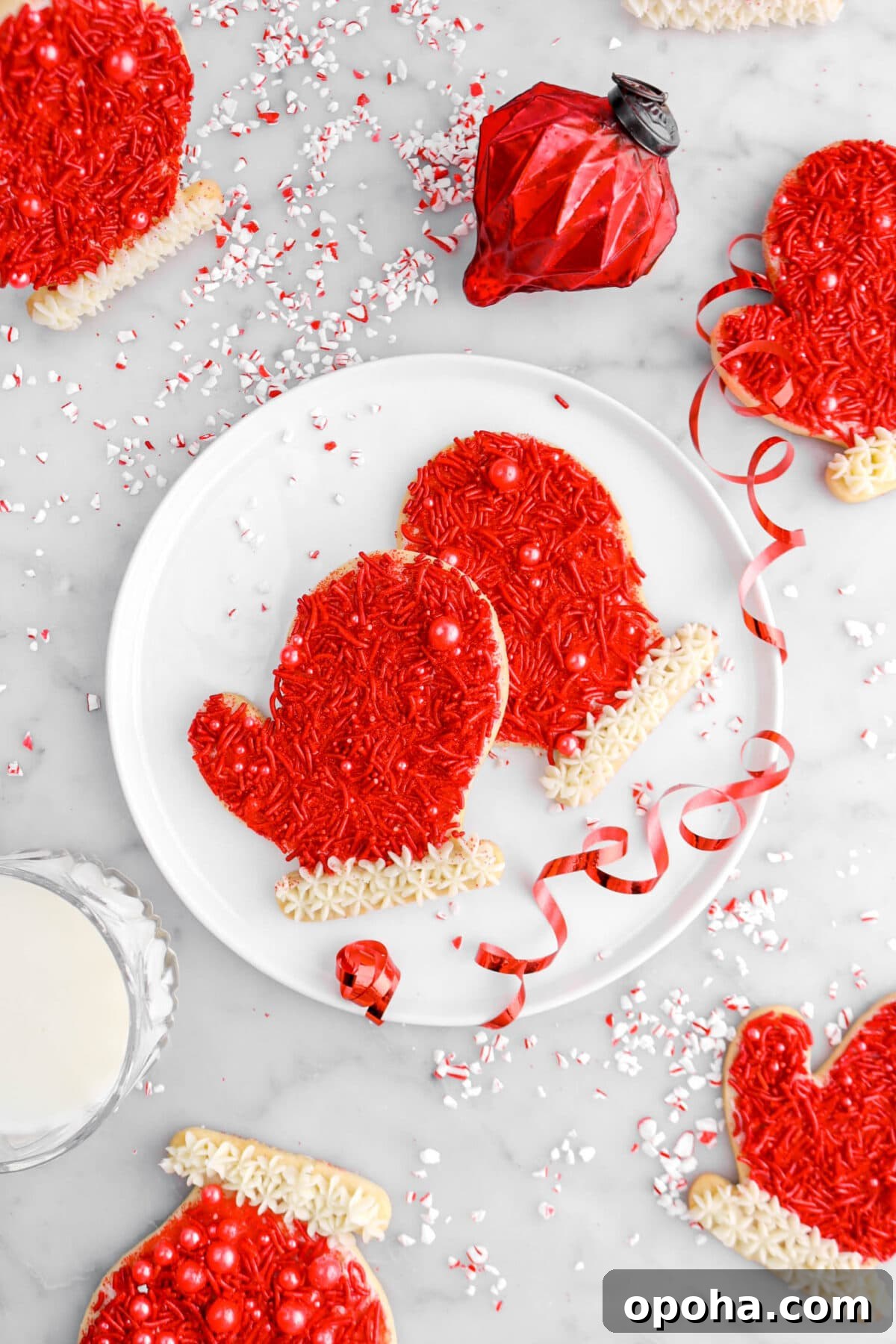 Santa’s Glove Peppermint Sugar Cookies: Festive Holiday Recipe 4 overhead shot of two glove cookies on white plate with red ribbon, red ornament, glass of milk, and crushed peppermint around.