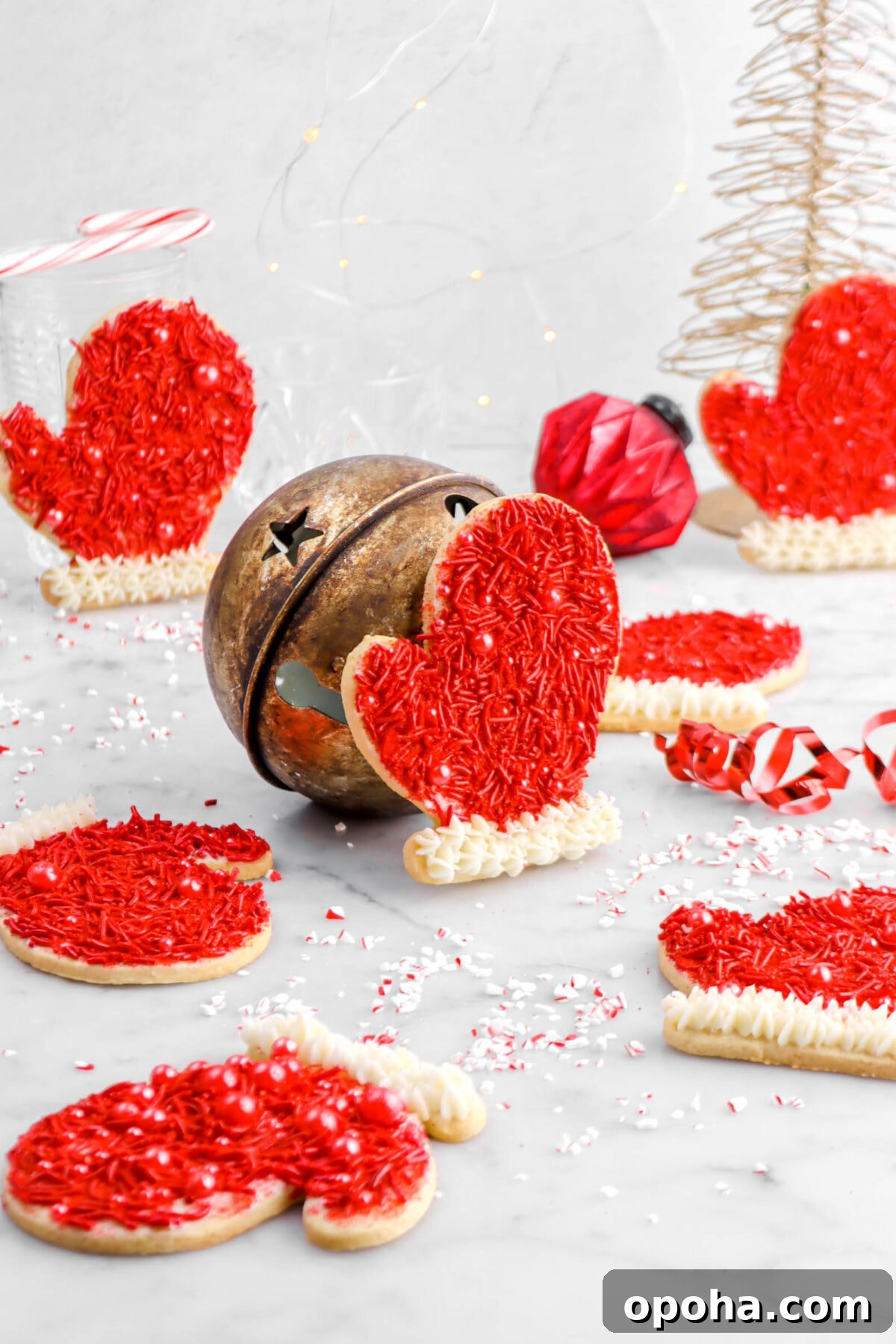 Santa’s Glove Peppermint Sugar Cookies: Festive Holiday Recipe 19 pulled back shot of seven glove cookies scattered around marble surface with one cookie leaning against a gold bell in the middle, another propped against a glass with a candy cane, and one leaning against a gold wire tree behind with red ornament beside.