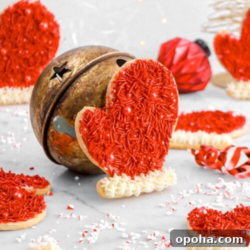 Santa’s Glove Peppermint Sugar Cookies: Festive Holiday Recipe 20 glove shaped sugar cookie propped against large bell, with six more cookies around, red ribbon beside, a red ornament, and fairy lights behind.